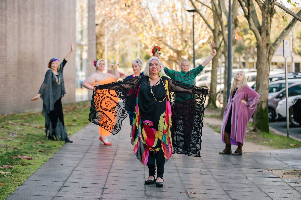 A woman stands in an outdoor area wearing a black dress with bright coloured parrots on it. Her arms are stretched out and she has a black lace shawl over them. Behind her is five people in fashion poses. One is wearing a long black and green dress, one is wearing a fluoro orange dress, one a blue outfit, one is in a green and black skivvy with maroon pants, and one in a velvet light purple dress. 