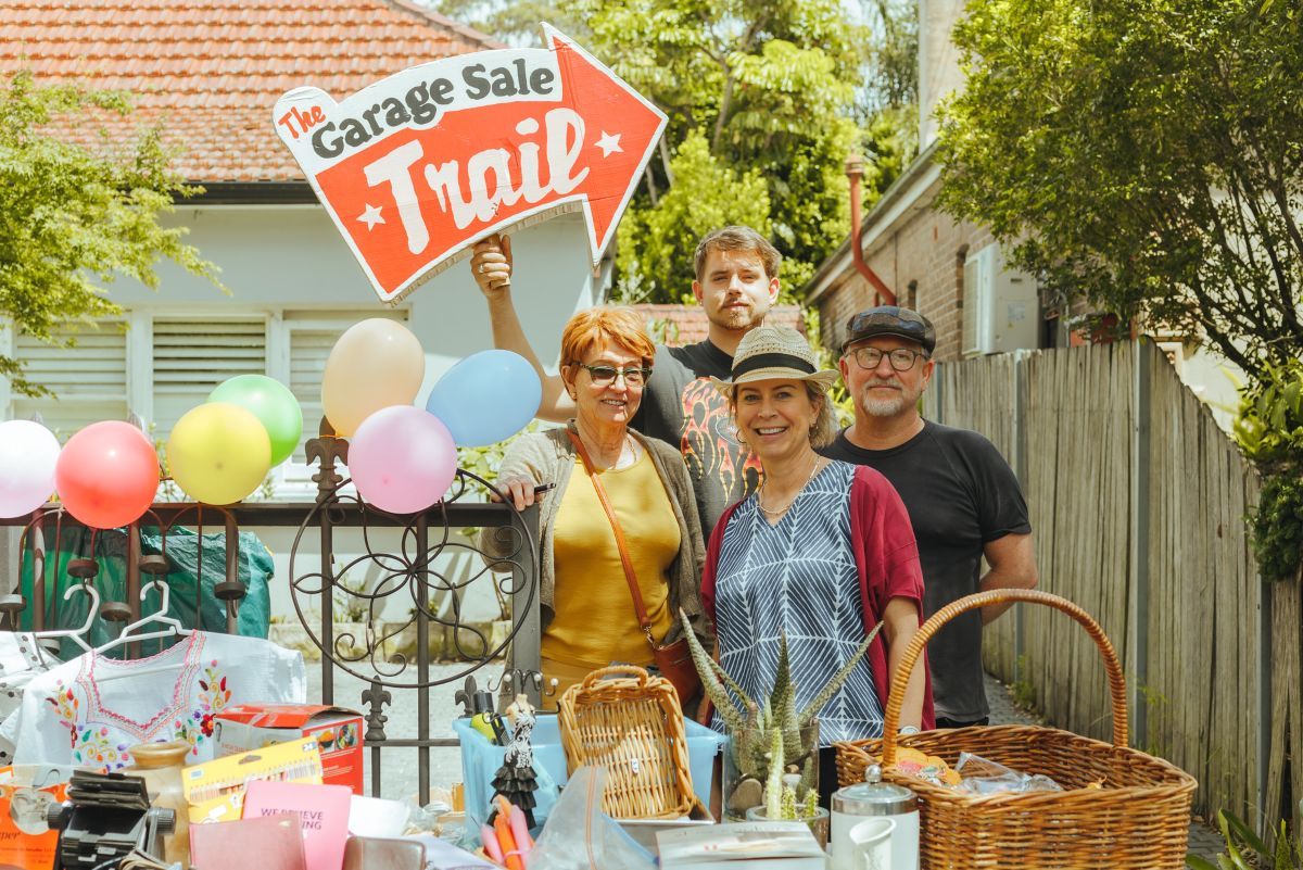 Two men and two women stand in a group smiling. There is a table in front of them filled with preloved items. One man at the back of the group is holding a sign that says 'The Garage Sale Trail' 