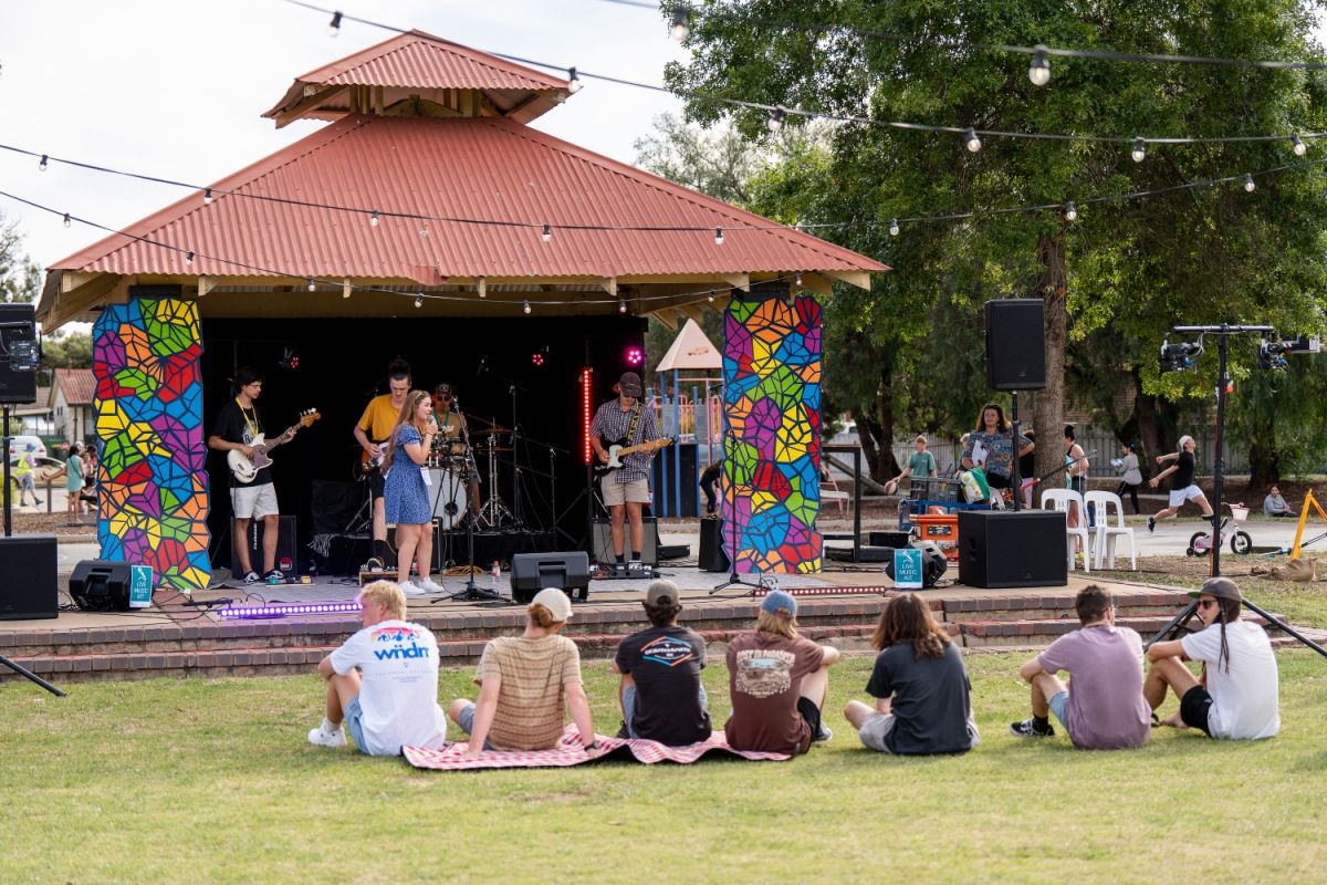 Wide shot of an outdoor stage in Chambers Park where a young, local band plays. A small crowd of young people are lounging on the grass in front of the stage.