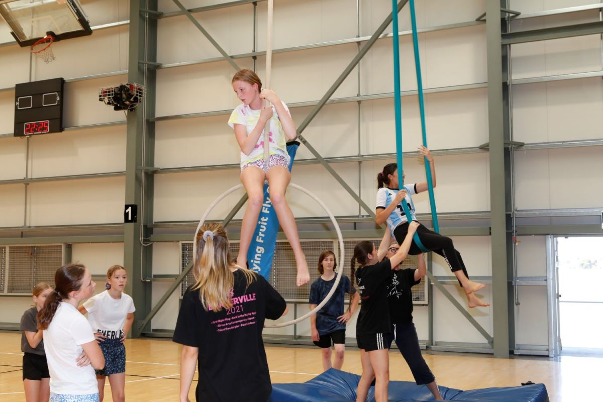 A crowd of young performers stand around an aerial hoop and a silk ribbon extending from the ceiling. Two trainers help youth sit atop the two obstacles.