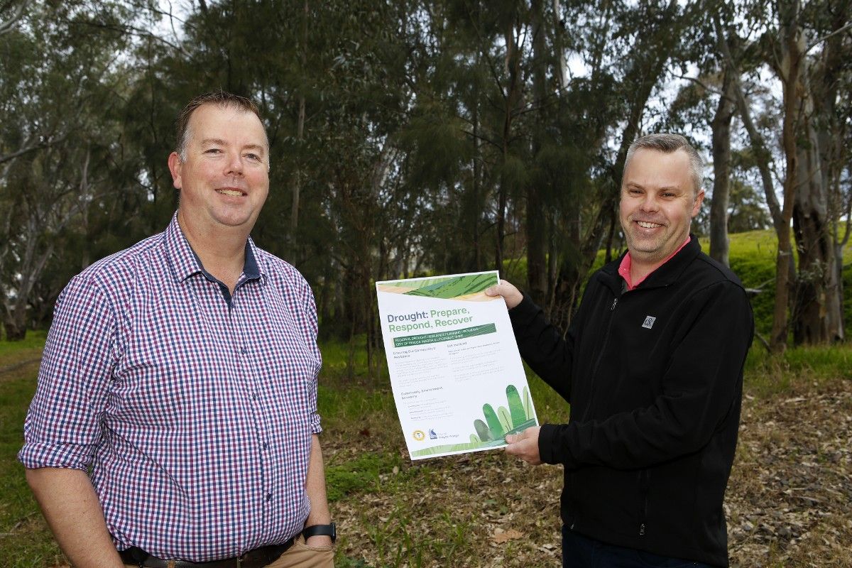 Two men standing in front of gum trees, with one man holding a poster about the Drought Resilience Plan