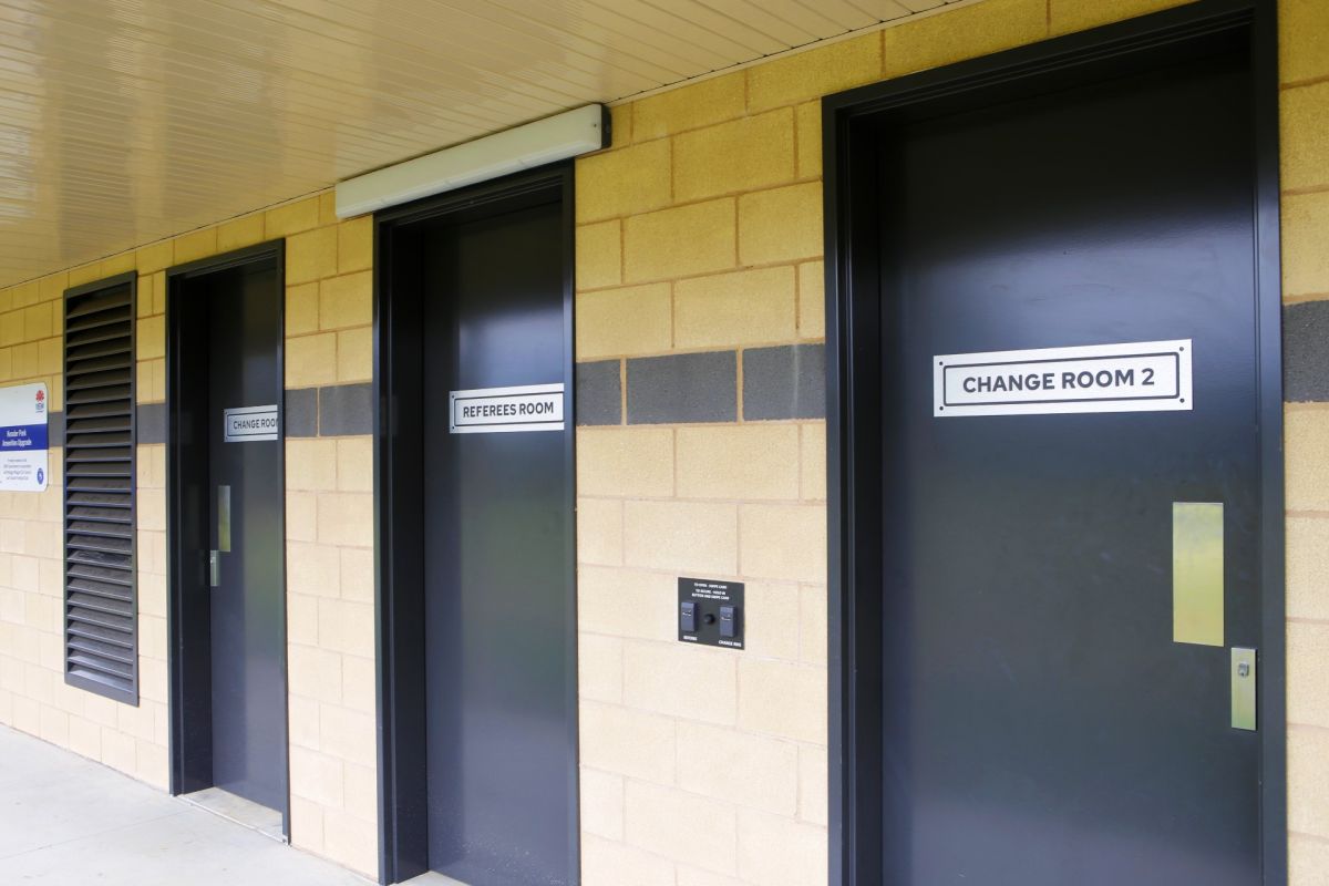 Three black doors on a brick building with signs on them indicating they are change rooms and a referees room, and a swipe card access panel between the doors.