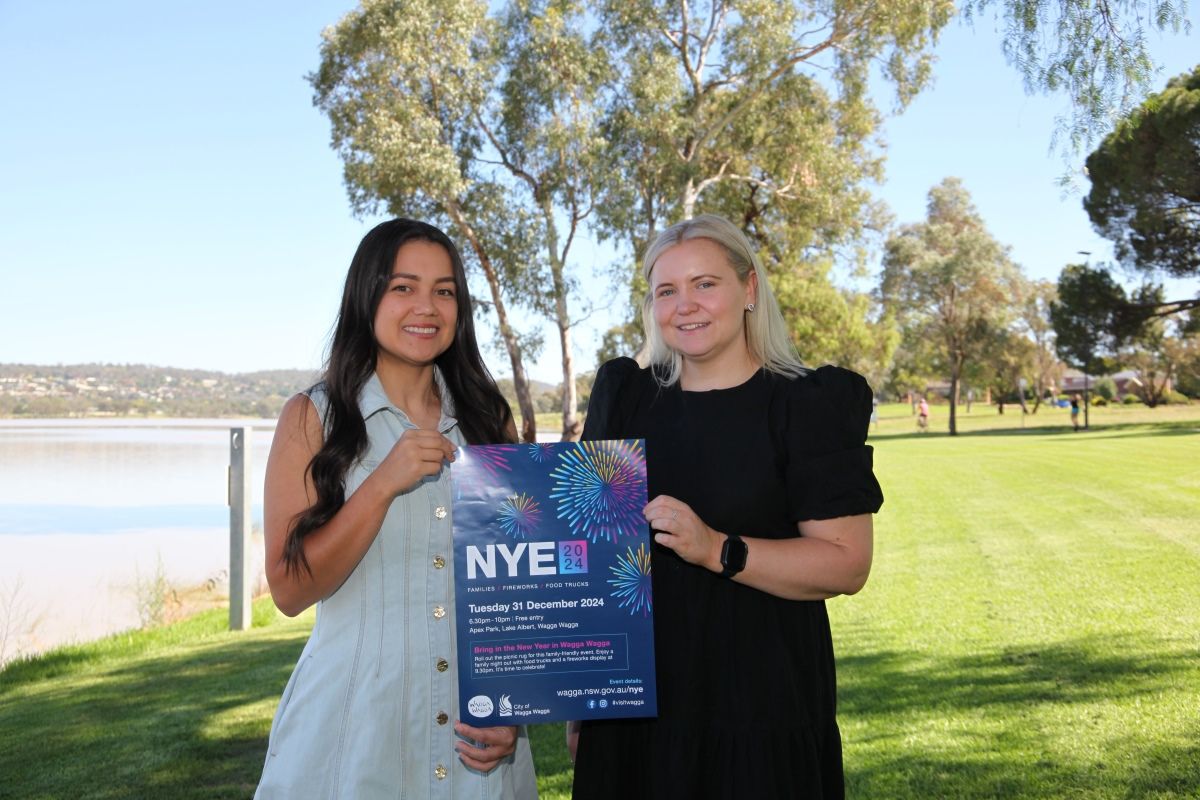 Two women holding a colourful poster saying NYE2024, with a grassy area and banks of a lake in the background