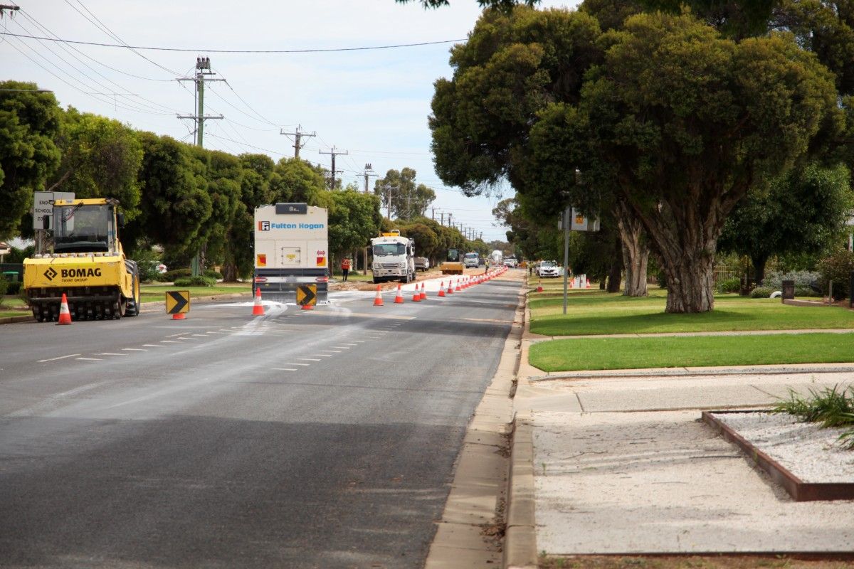 Roadworks underway on suburban street, with a row of witches hats stretching into the distance marking off the area where heavy roadworks machinery and trucks are carrying out the roadworks.