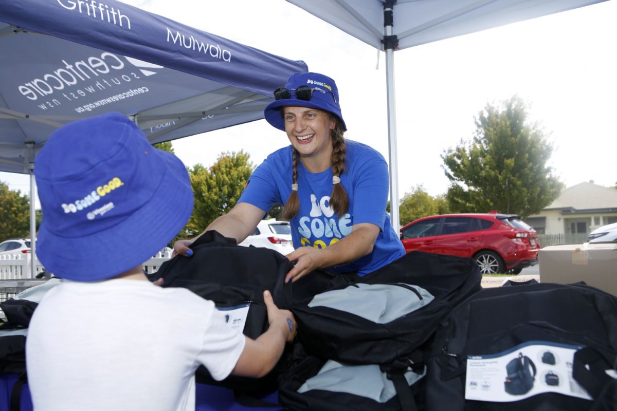 Woman wearing a blue shirt with Do Some Good printed on it hands over a black school backpack to a young boy.