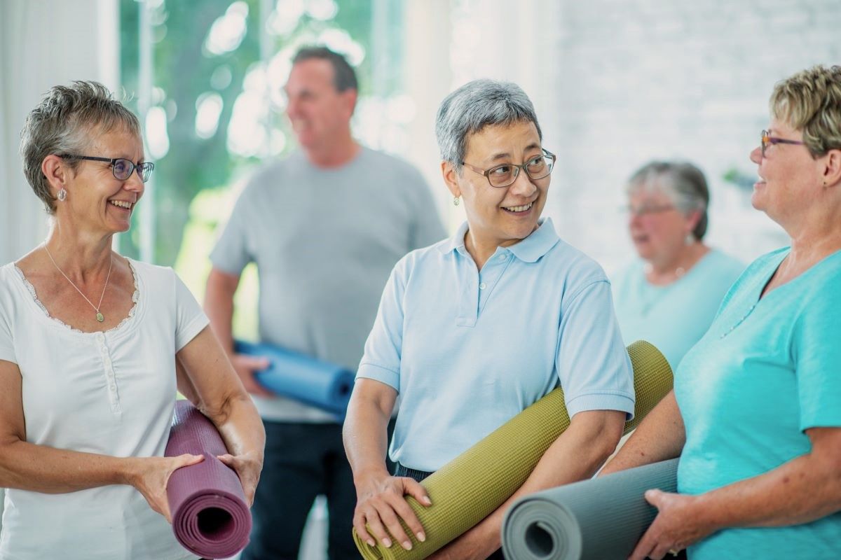 Five older people in casual dress and holding yoga mats, smiling at each other.