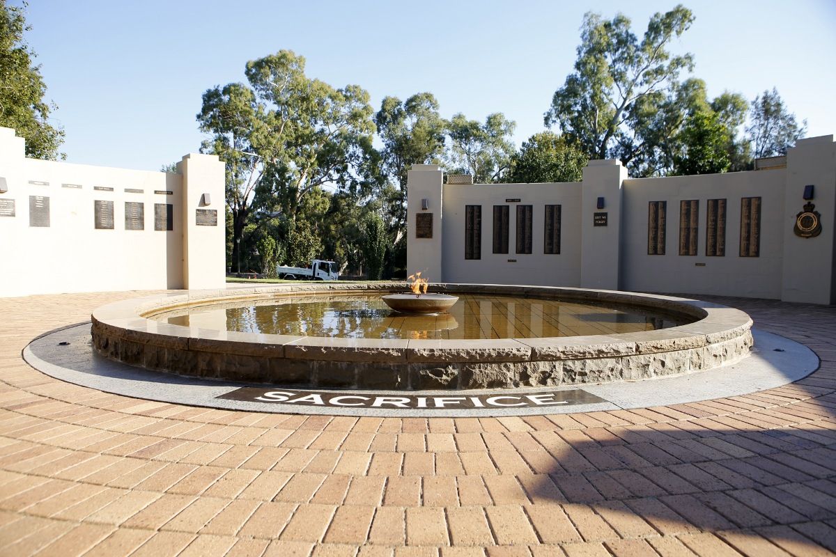 Eternal flame inside bowl set in pond, with the word sacrifice engraved in a stone paver next to the pond and Honour Rolls on the walls of a memorial in the background.