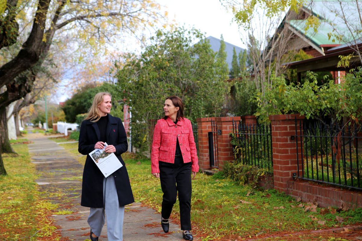 Alt text: Two women walk along a quiet, tree-lined suburban street on a crisp autumn day. The woman on the left, wearing a long black coat and light grey trousers, holds a housing strategy document, and smiles as she speaks. The woman on the right, dressed in a red textured jacket and black pants, listens attentively. They walk past brick fences and green foliage, with fallen yellow leaves scattered along the footpath.