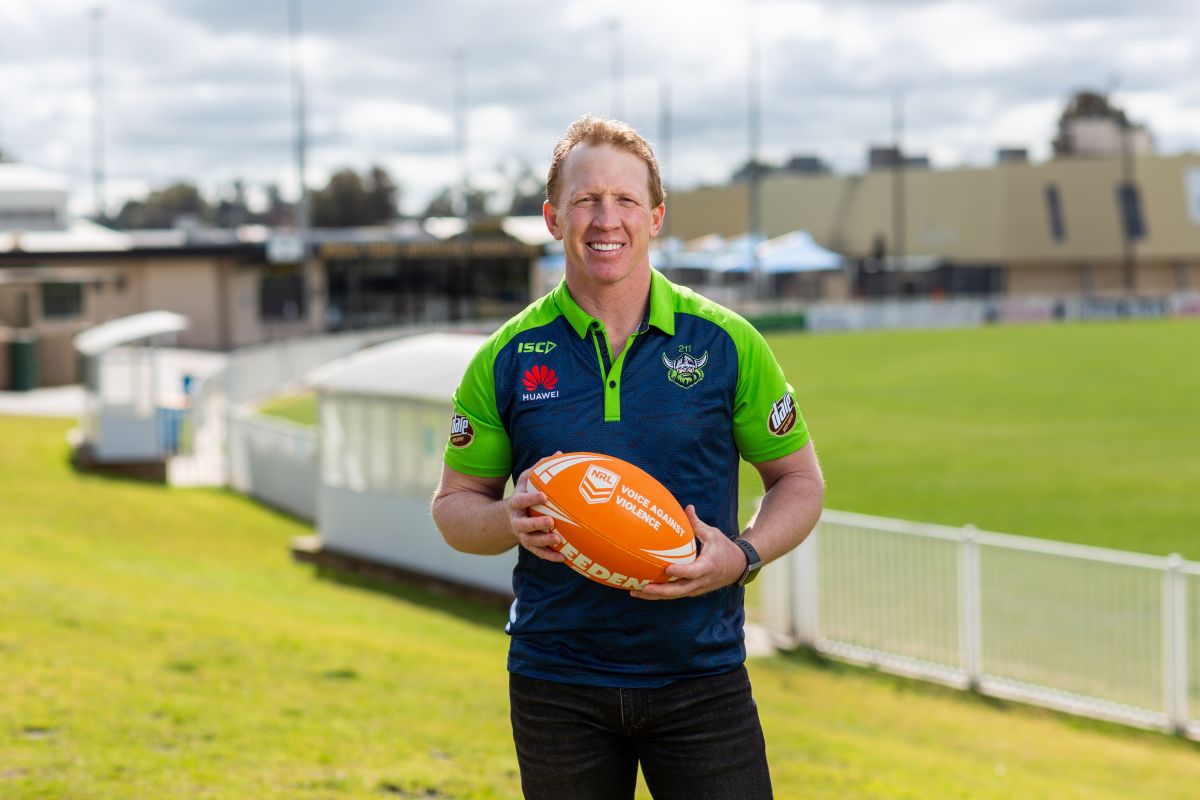 Man standing at football ground holding a rugby league ball with Voice Against Violence on the ball