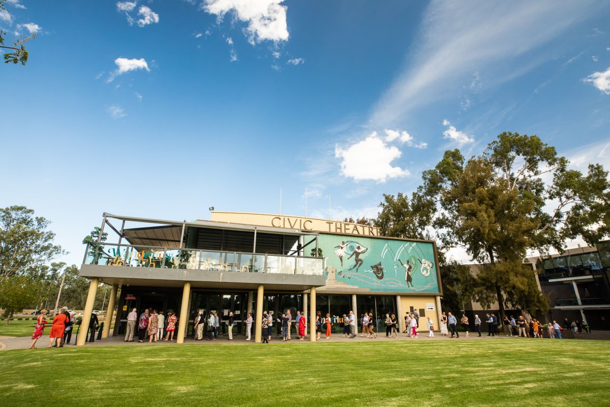 People lined up outside theatre, with blue sky in background and grass in foreground
