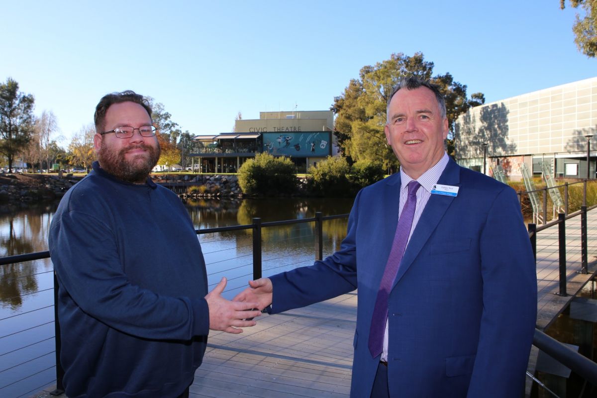 Two men shaking hands, standing on boardwalk next to Lagoon with Council building in background