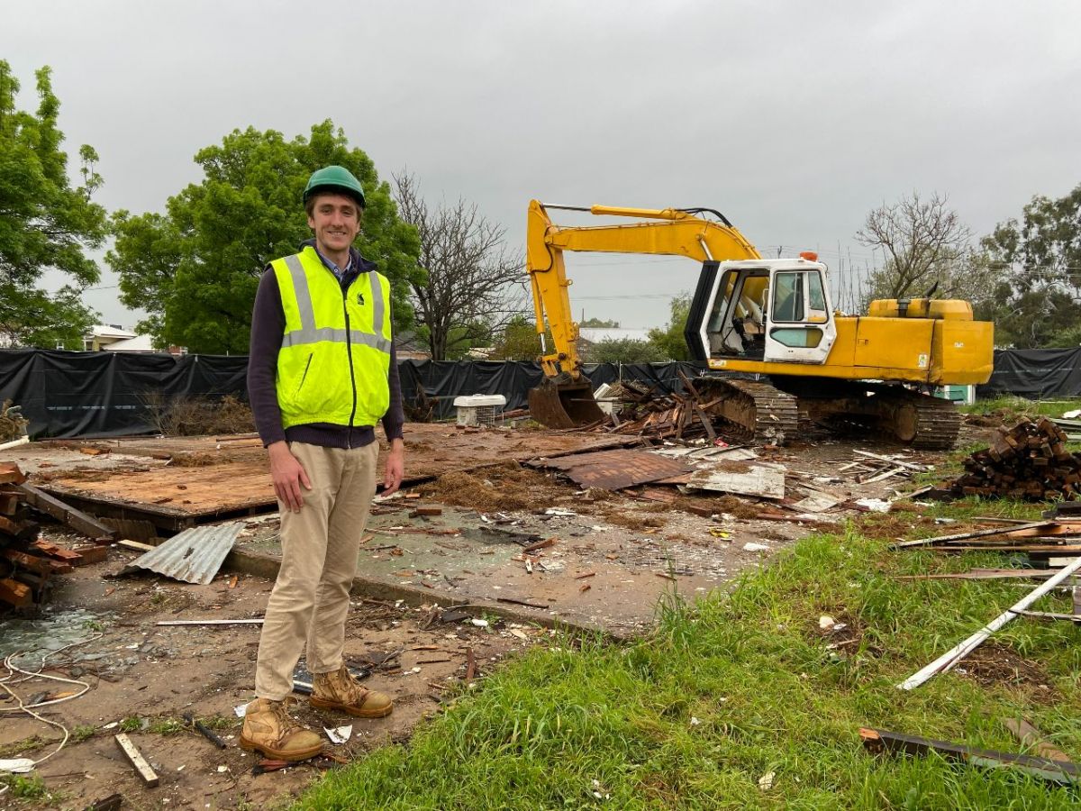 Man wearing hard hat and hi vis standing on worksite with excavator in background.