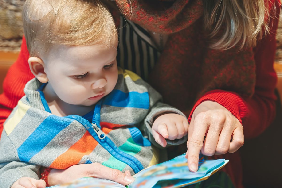 A baby sitting in its mother's lap points at a book.