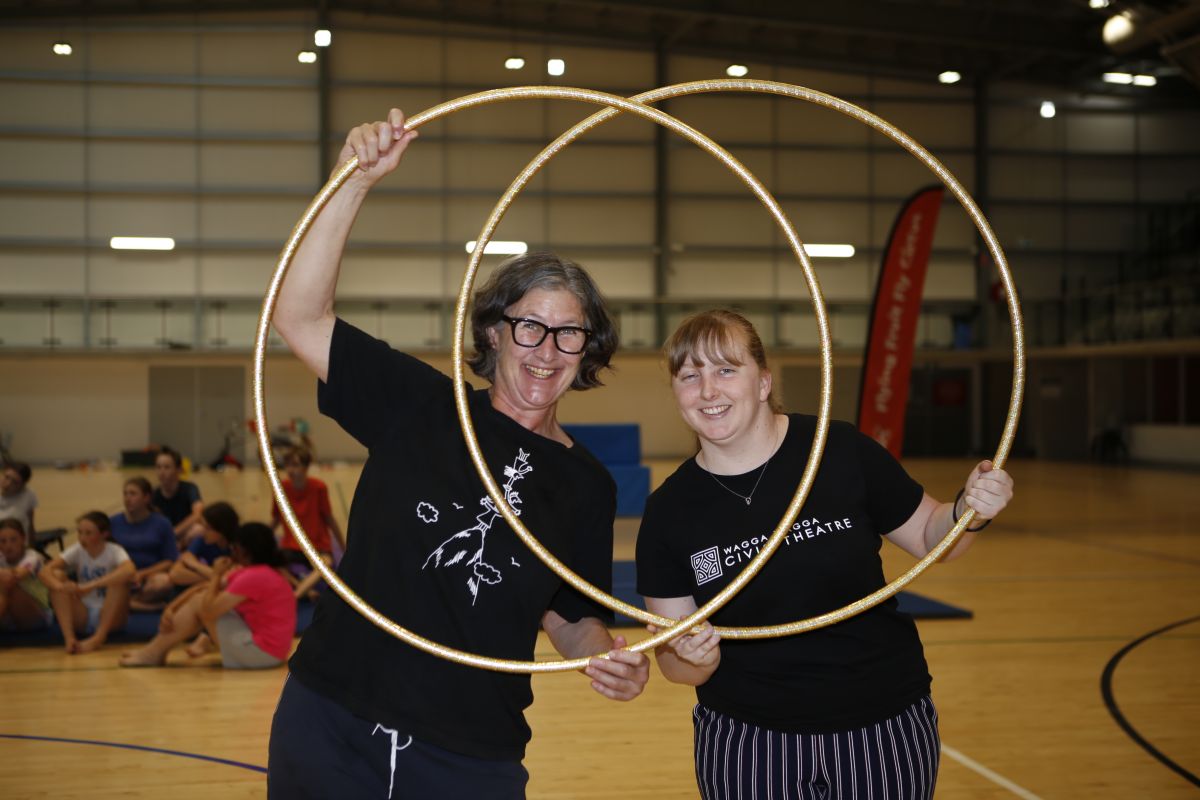 Two workers hold hula hoops together and peer inside to camera through the circle. Students from the camp can be seen in the background gathered around a large gym crash mat.