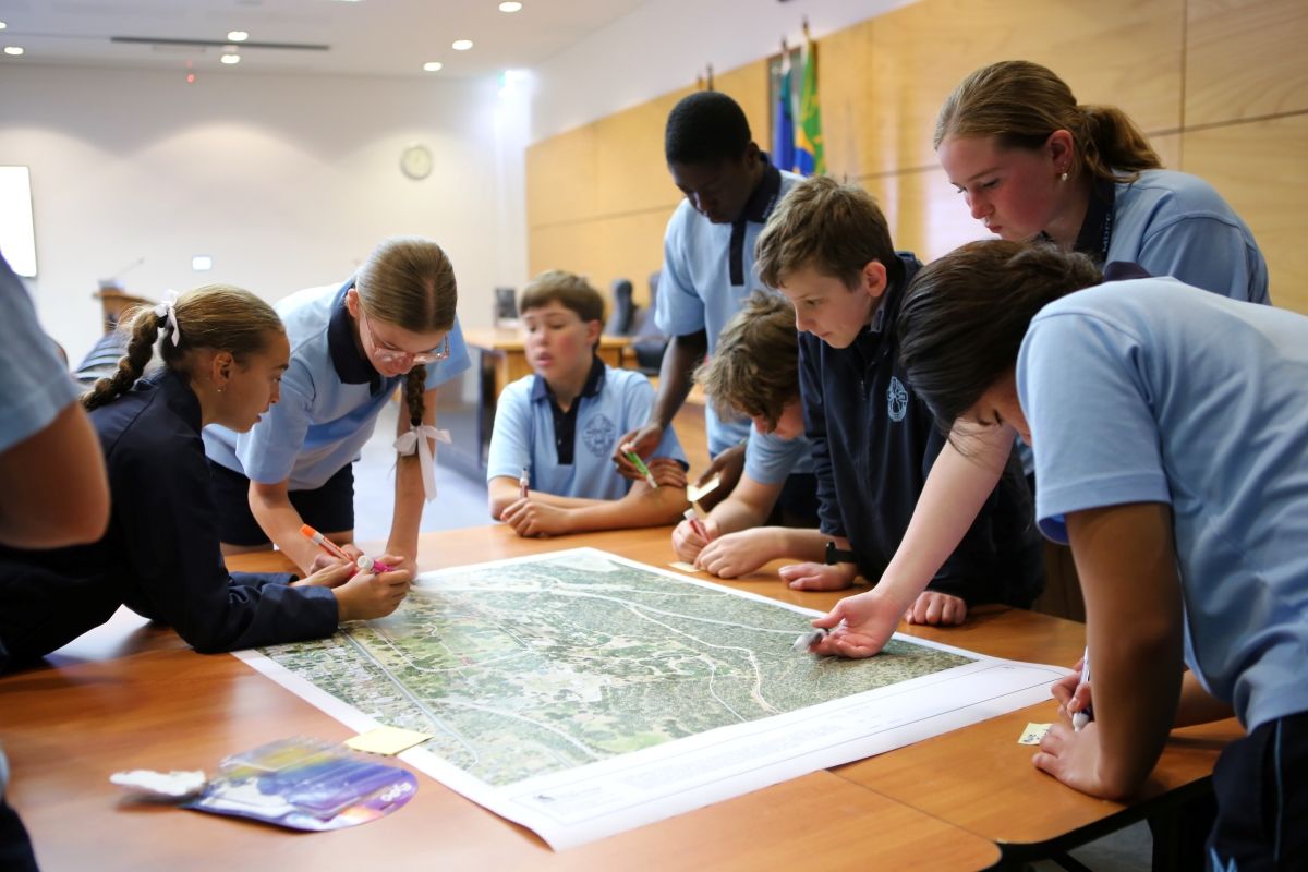 A group of young people, dressed in school uniforms, are gathered around a table with a large, printed map. Some students hold markers, others are visibly writing.