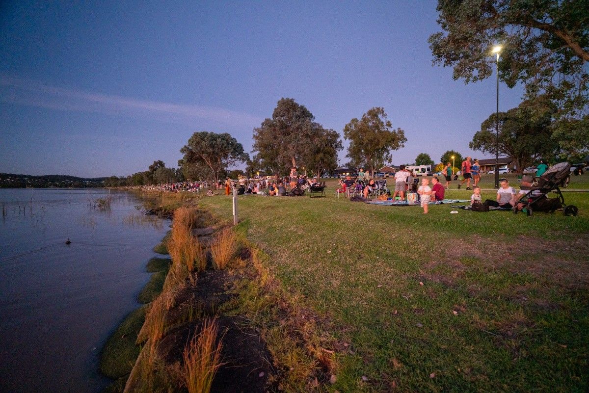 People and families sitting on picnic rugs or camp chairs on the grassed foreshores of a lake in the early evening.