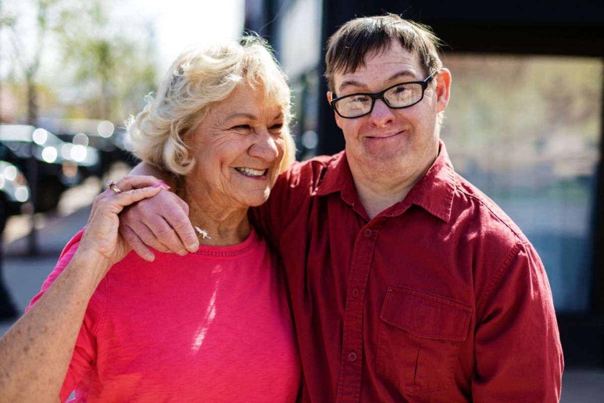 : Older woman and older man with Down Syndrome standing in a street in a shopping area, smiling.