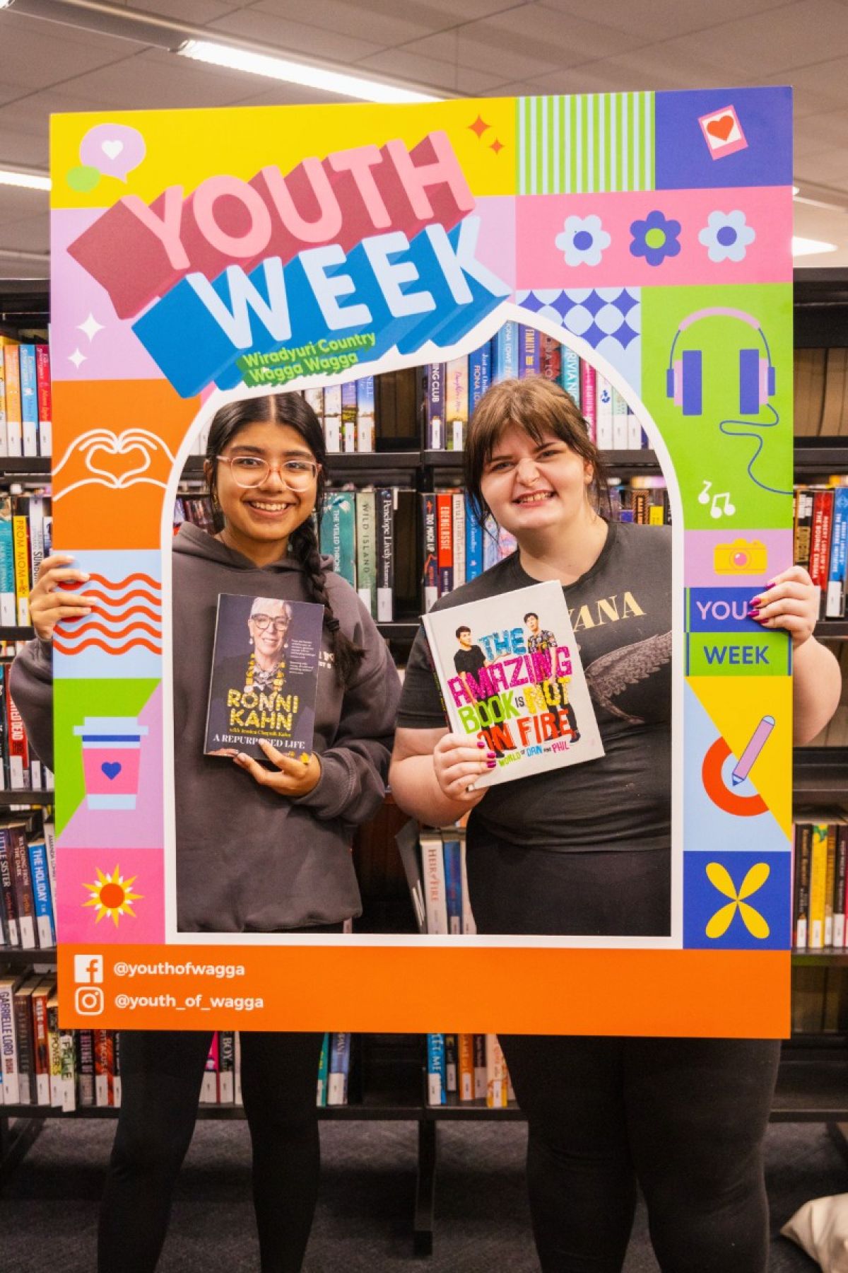 Two young people posing for photo inside frame that says Youth Week, with library books on shelves in background.