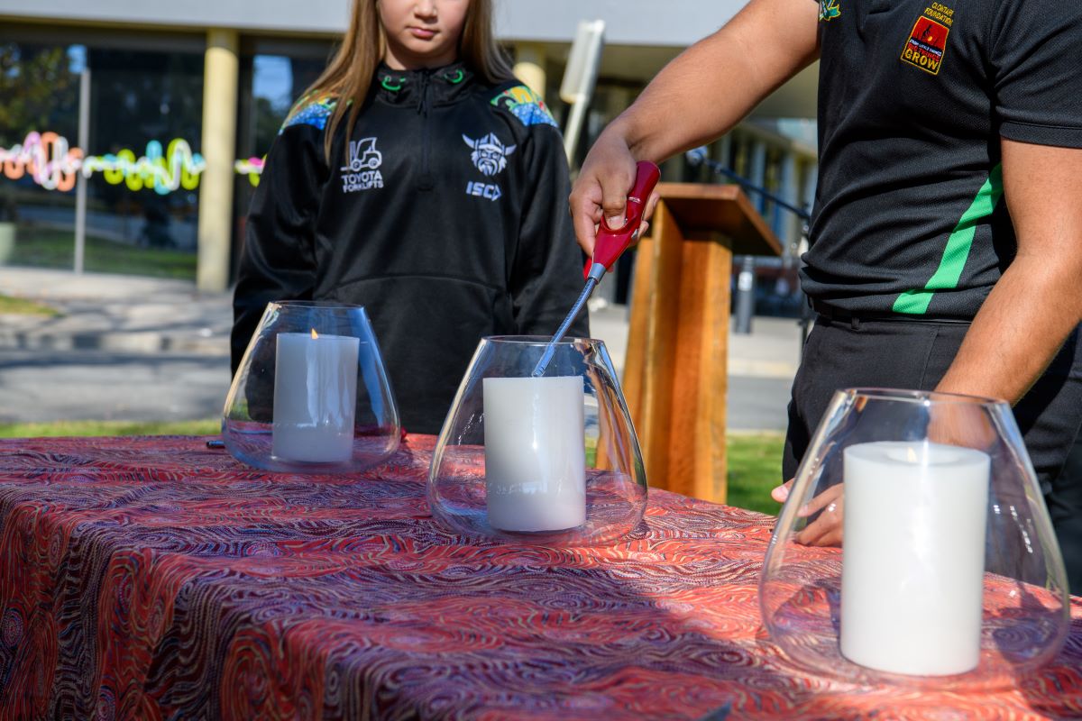 Two young people light candles in large glass jars at an outdoor event.