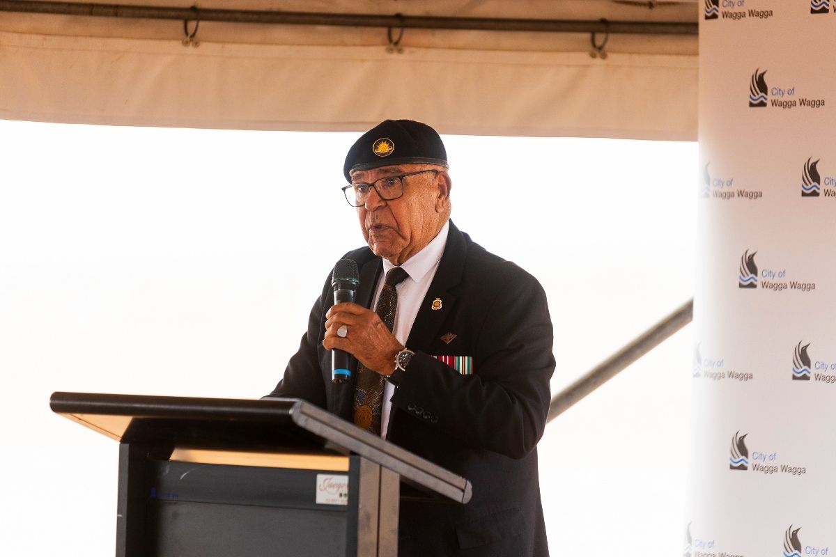 A First Nations man wearing a black beret with a gold Australian Army crest embroidered on the front, stands holding a microphone behind a lectern, as he gives a speech. His facial expression as he speaks, is peaceful, sincere and engaged