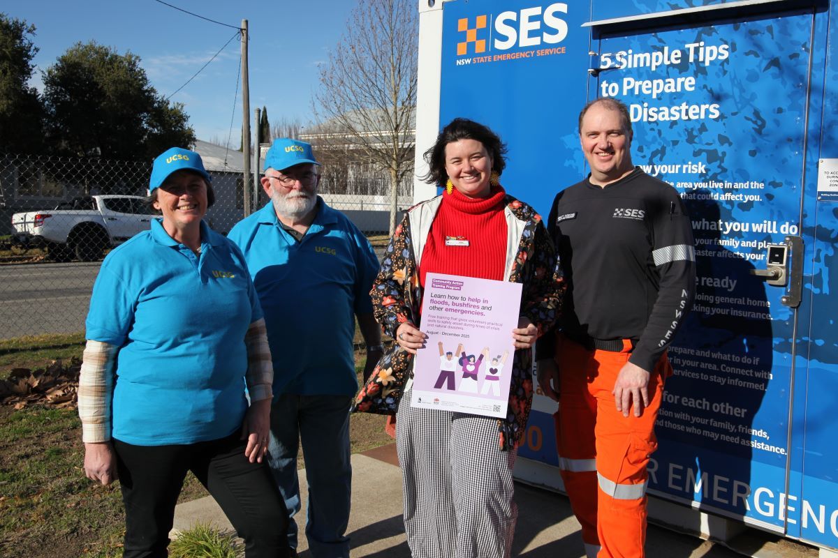 A man and a woman wearing blue shirts and hats, a young woman holding a poster, and a man wearing an SES uniform standing outside near a Community Assistance Team shipping container.